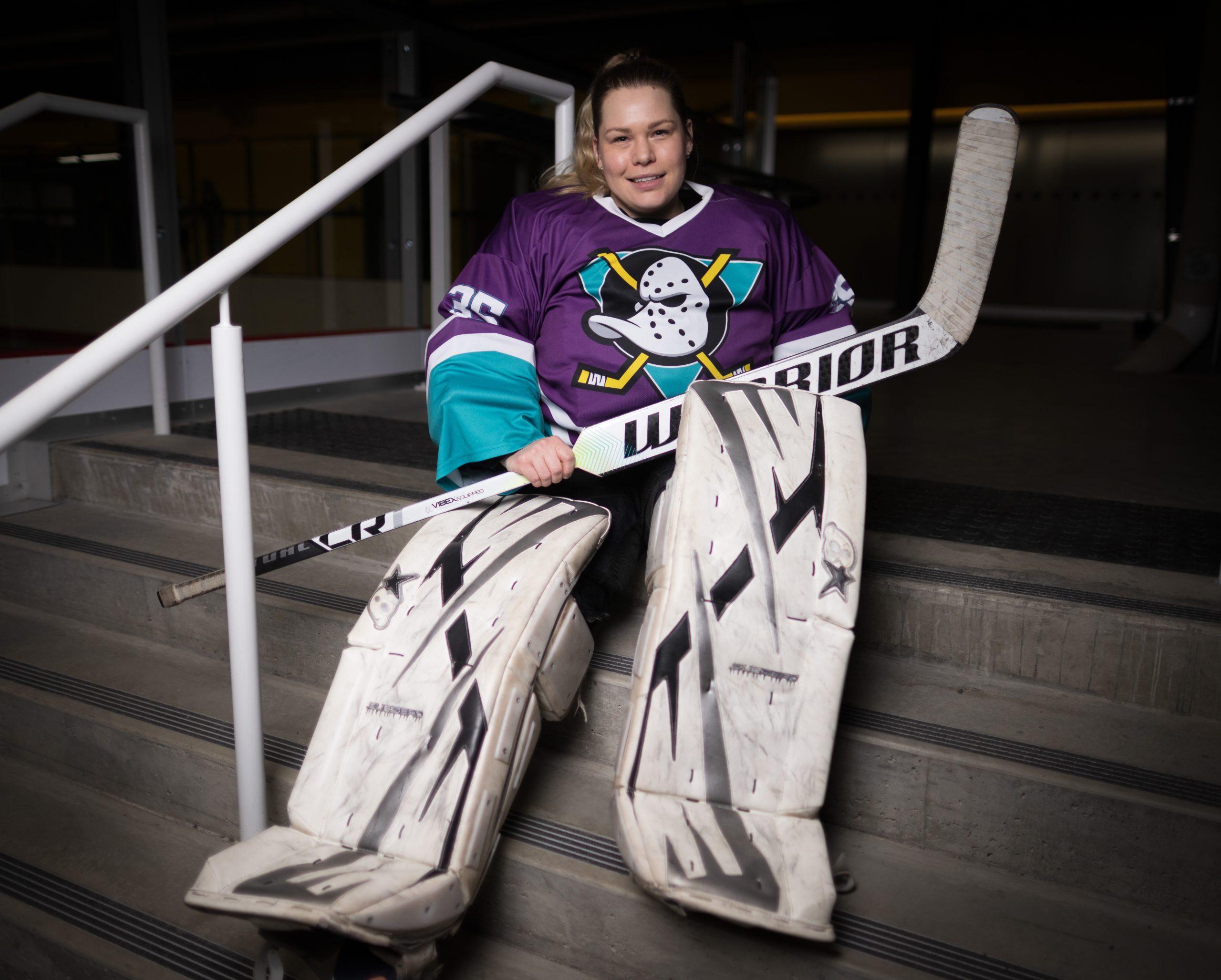 A woman in ball hockey goalie equipment sits on stairs in an arena.