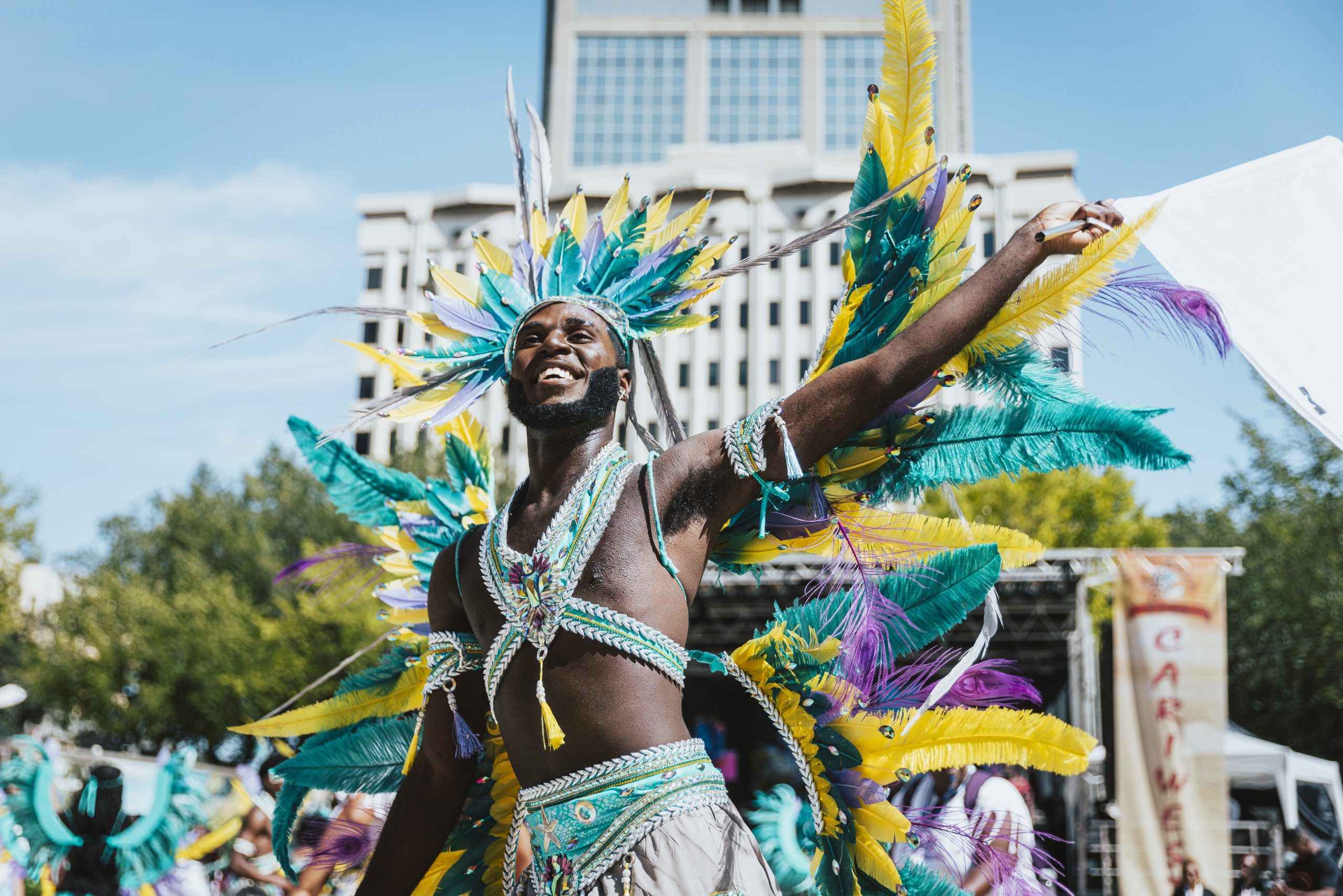 A Black man wearing a headdress and arm bands with yellow, purple and green feathers dances in front of a stage with a Cariwest banner handing from the side of it.