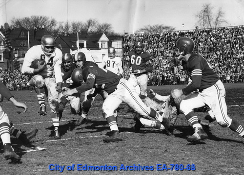 Football players in 1950s uniforms try to tackle a rival who is running with the ball tucked under his right arm.