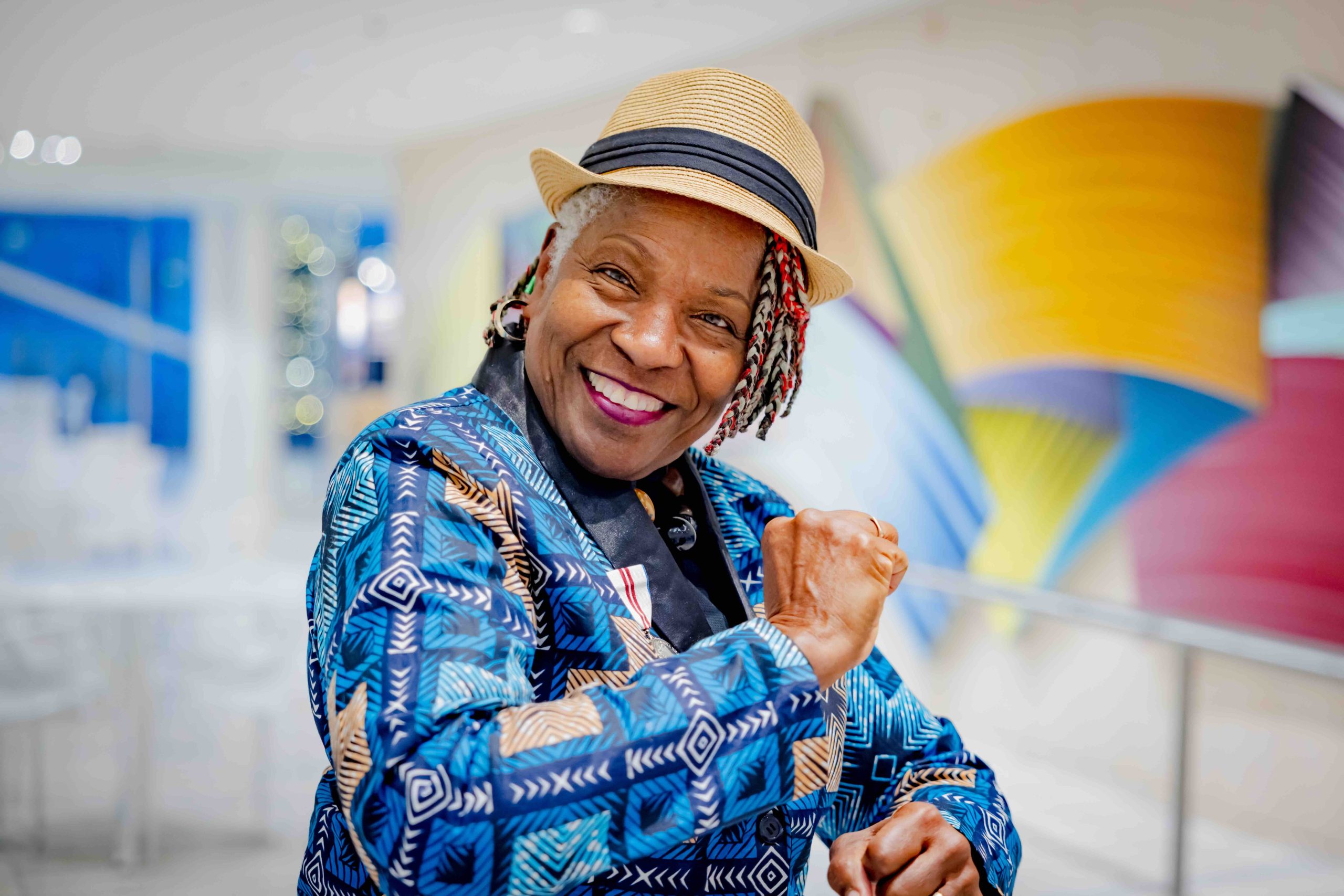 A Black woman, wearing a multi-patterned blue jacket and straw fedora, smiles at the camera as she makes fists with her hands.