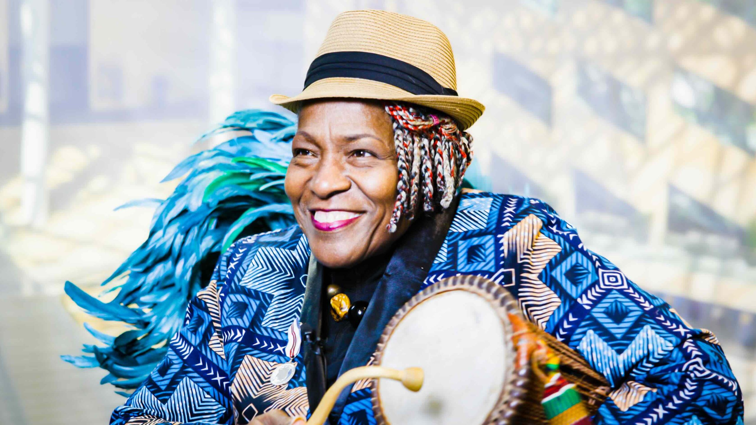 A Black woman, wearing a multi-patterned blue jacket and straw fedora, holds a drum under her left arm and a curved drum stick in her right hand.