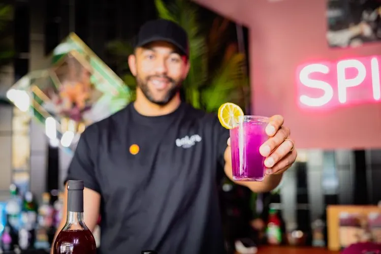 Keenan Pascal wearing a black Token Bitters shirt, smiles while holding up a vibrant purple mocktail with lemon garnish. The background features a neon sign with the word "SPILT".