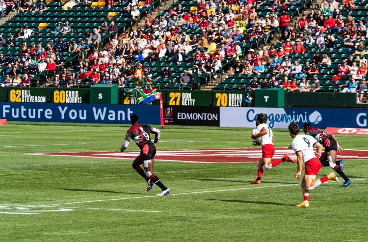 Two international rugby teams compete on a field in front of fans in the stands on a sunny day.