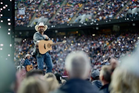 Garth Brooks, holding a guitar, stands on a stage surrounded by fans on both sides.