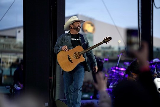 Garth Brooks smiles and he walks on stage as he holds a guitar.