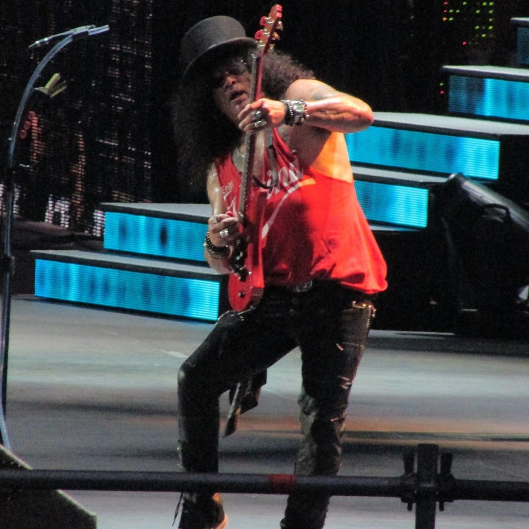 Slash, a guitarist with Guns N' Roses, wears a black hat and sunglasses as he leans back and plays his guitar.