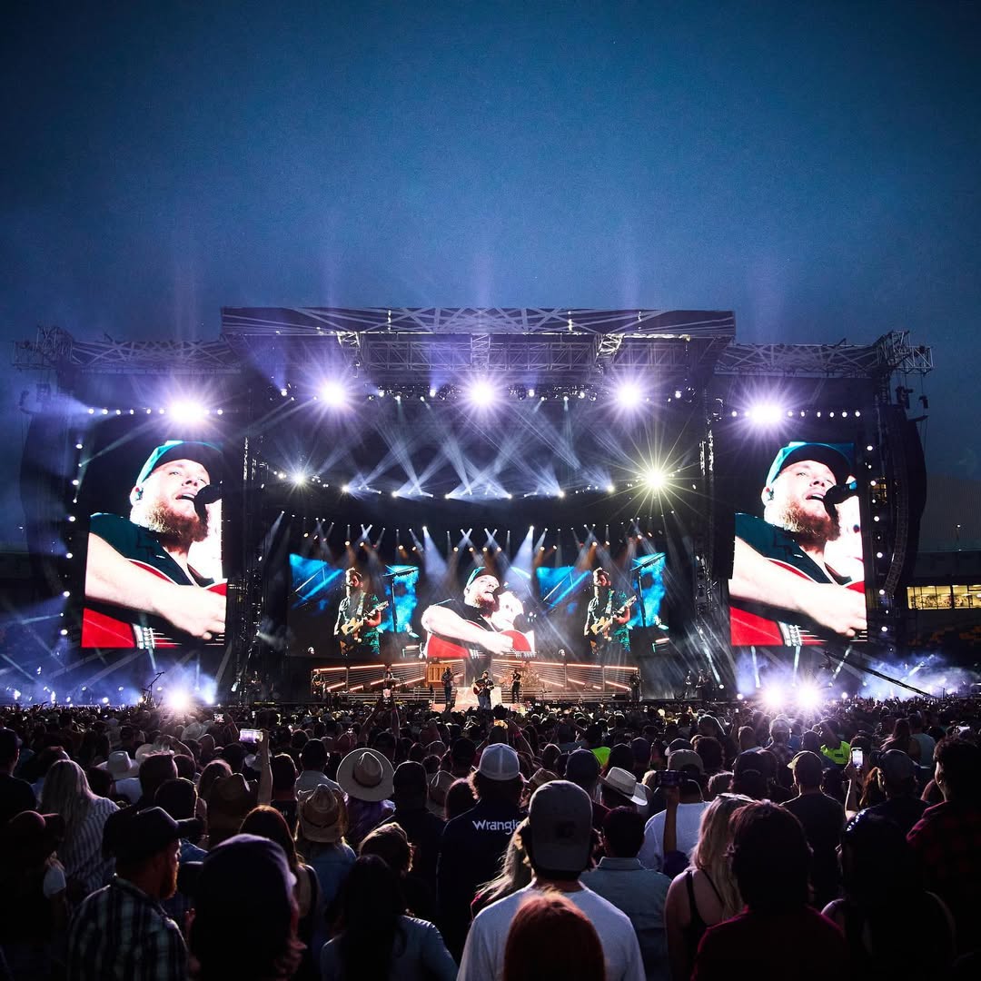A shot of the backs of fans standing in front of Luke Comb's stage, which includes three large TV screens featuring Combs singing into a microphone and playing a guitar.