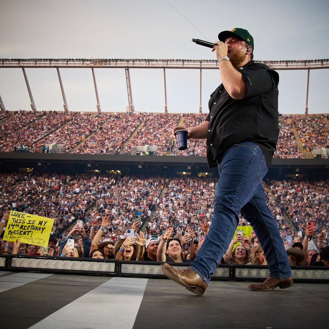 Singer Luke Combs holds a plastic cup in his left hand and sings into a microphone in his right hand as he walks along a stage in front of fans cheering and taking photos with their cell phones.