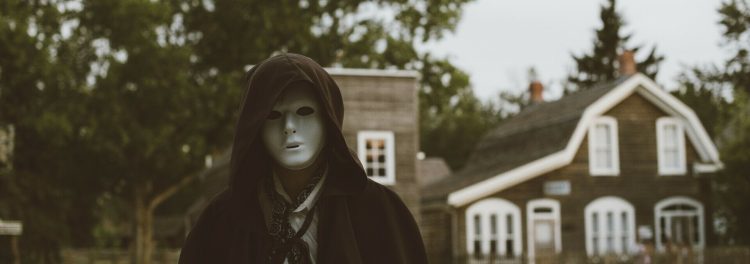 A person wearing a white mask and a black hooded cape in front of historic buildings. Credit: Fort Edmonton Park