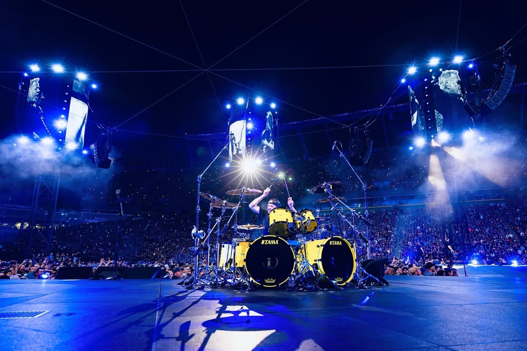 Lars Ulrich sits behind and plays a drum kit on a stage surrounded by thousands of people.
