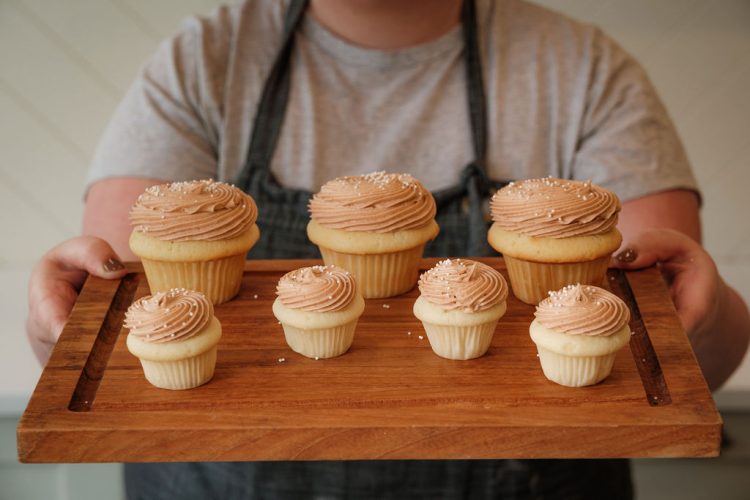 Various cupcakes with swirled icing on a tray held up by a torso wearing an apron.