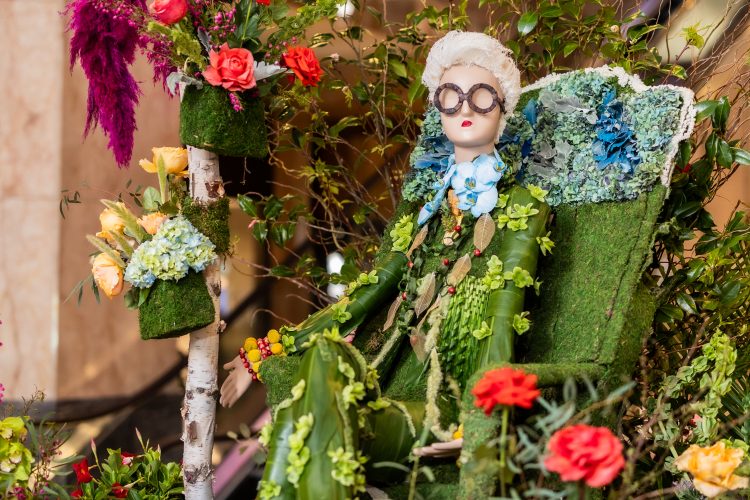 A mannequin with a white short wig, sitting in a chair with her clothes and chair covered of flowers and greenery.