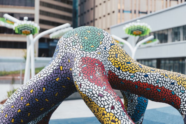 A sculpture that resembles a floating octopus with five tentacles and multi-coloured mosaic tiles stands outside a few buildings.
