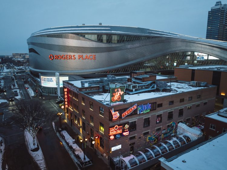 Aerial of a large silver arena behind a brick building with several neon signs attached at dusk in winter