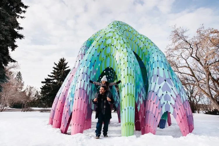 A young girl sits on her father’s shoulders as the two walk through a large public art sculpture in a snow-covered park on a cloudy winter day.