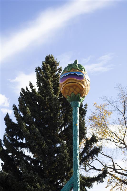 A large, bright yellow pineapple sculpture with sunglasses stands guard over a playground with swings, monkey bars, and drums.