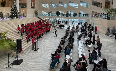 A children’s choir dressed in red performs on stairs in front of a crowd seated and standing around a large room.