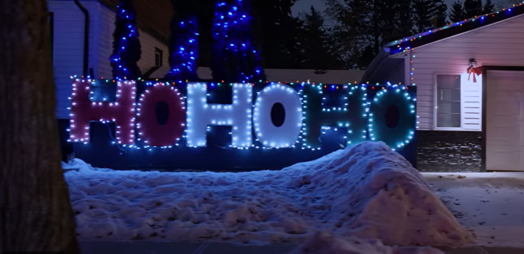 A giant red white & green lit up HOHOHO sign sits on a snowy yard next to a house.