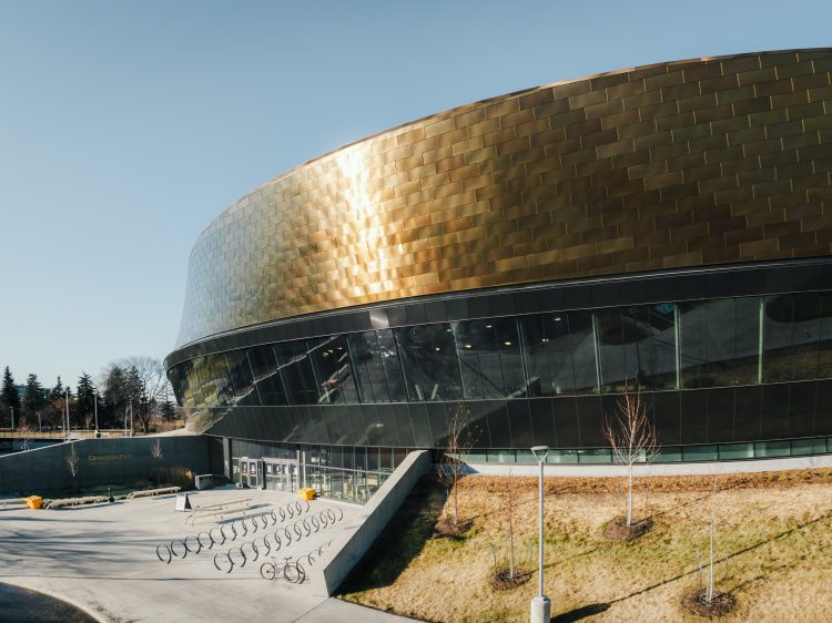 The main entrance of a round, copper-tiled recreation centre with large windows on a sunny day.