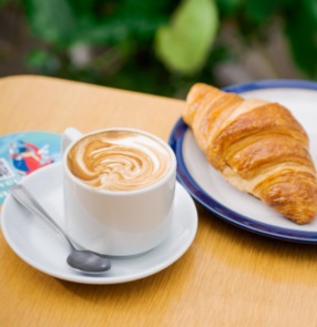 A latte with swirled milk and a croissant on plates on a coffee table. CR: OSBA