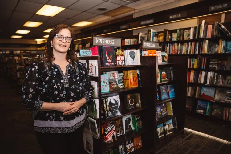 A woman wearing glasses stands next to bookshelves with the sign "New Releases" on top of them.