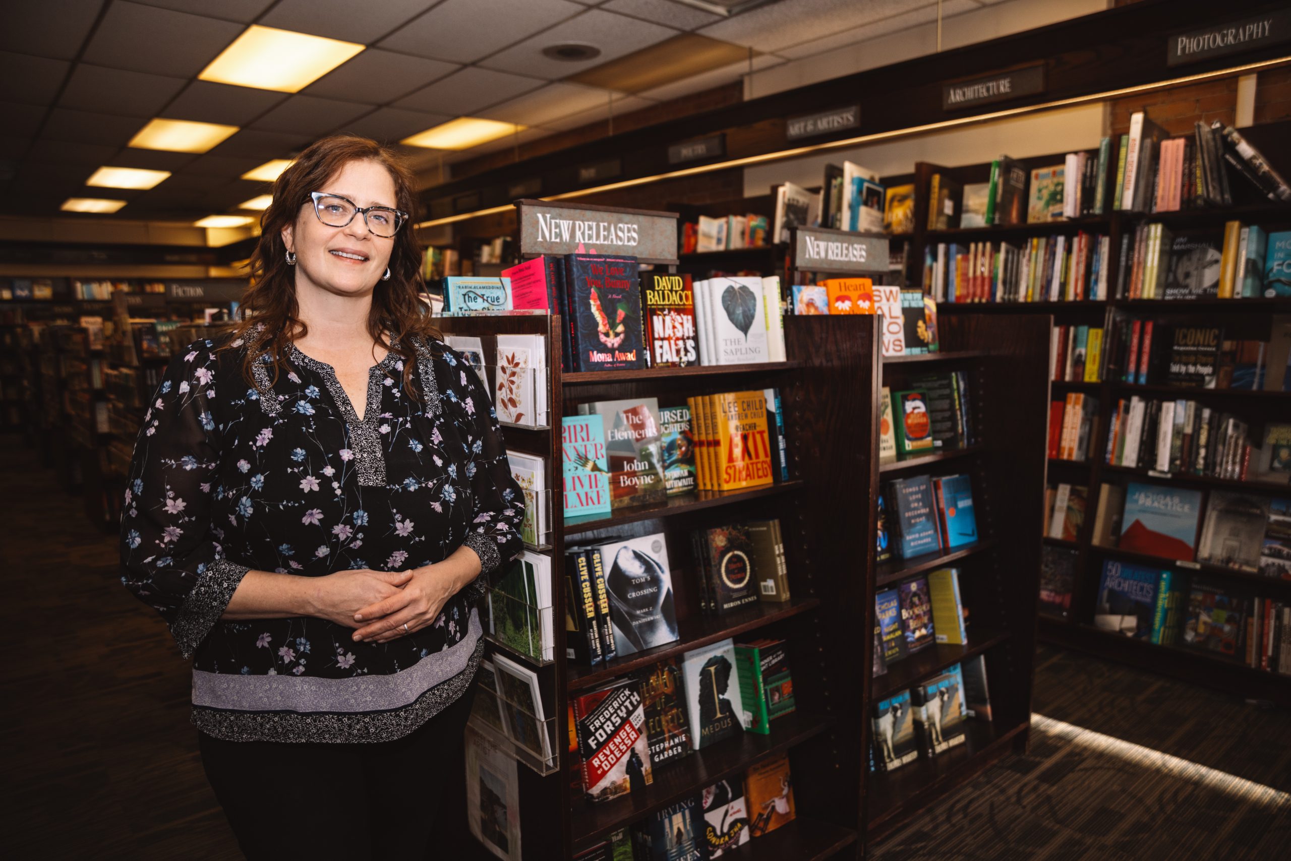 A woman wearing glasses stands next to bookshelves with the sign 
