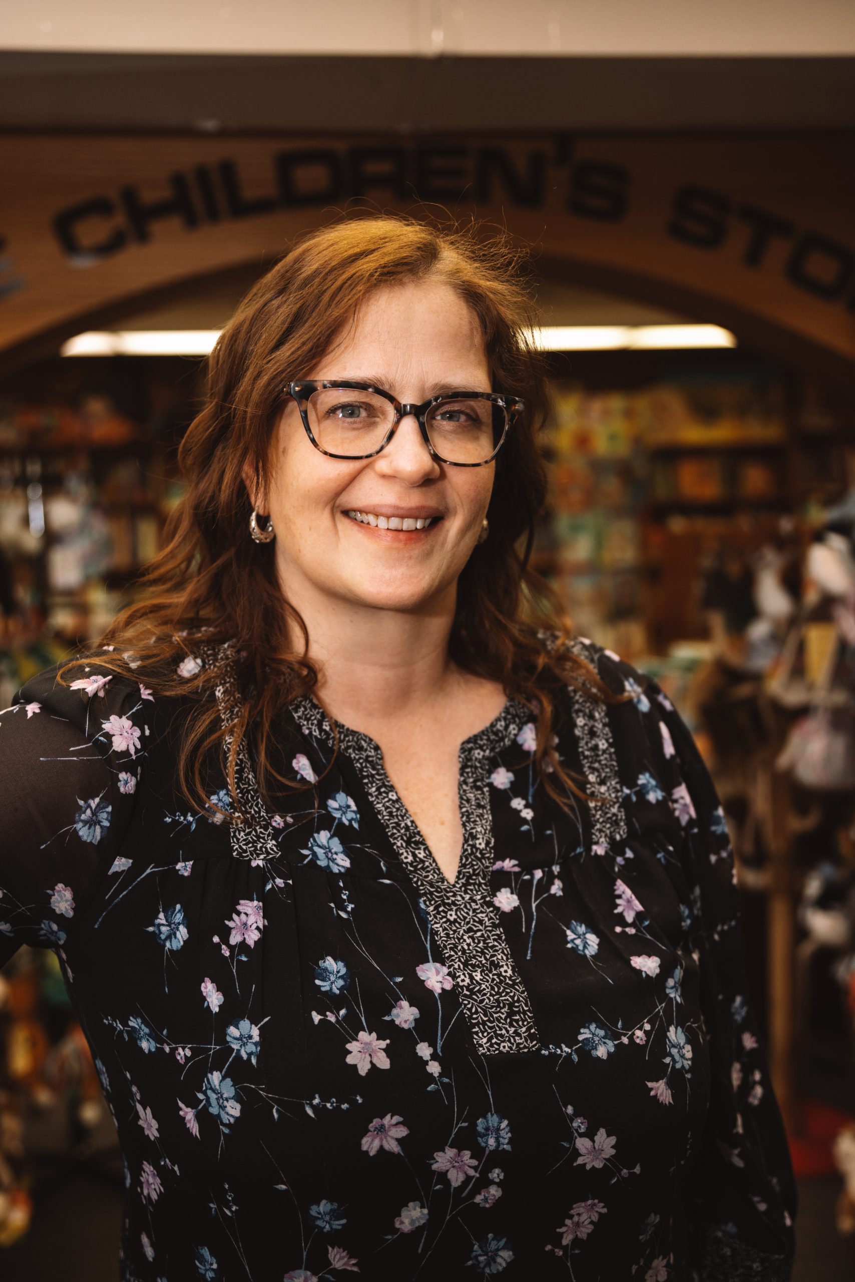 A woman, wearing glasses, smiles as she stands in a store full of bookshelves.