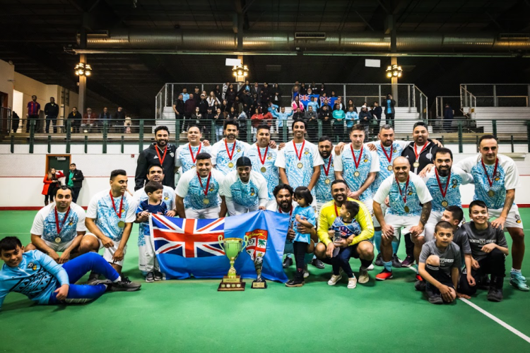 A men’s soccer team representing Fiji poses together wearing medals, with a trophy and flag.