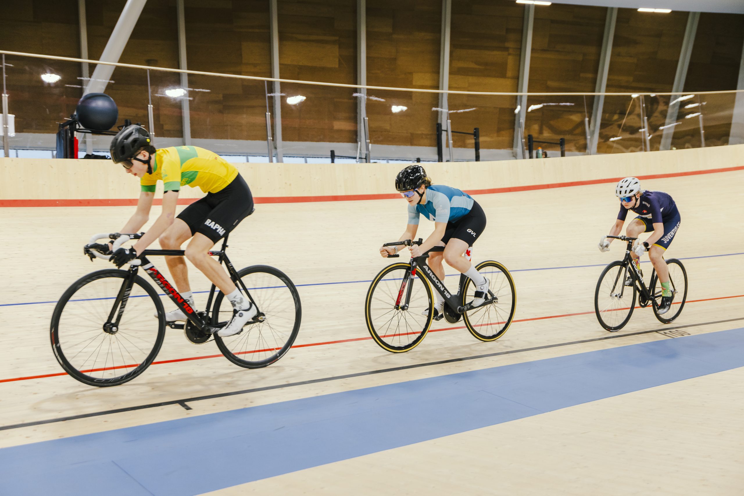 Three cyclists, in single file, race on the wooden track of an indoor velodrome.
