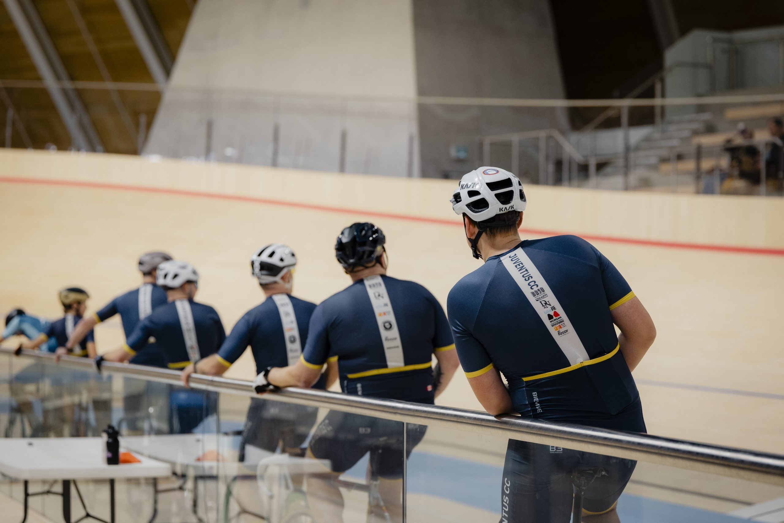 Seven cyclists, including six in Juventus Cycling Club bike jerseys and shorts, sit on their bikes and rest their arms on the silver railing circling the interior perimeter of an indoor cycling track.