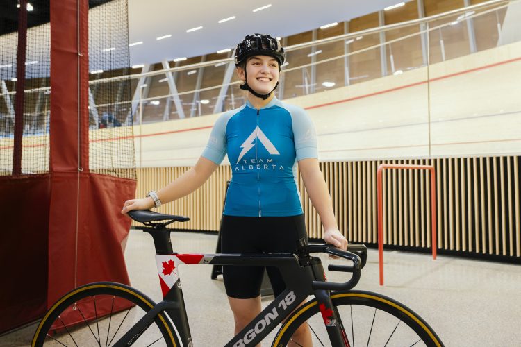 A teen girl, wearing a helmet, a Team Alberta bike jersey and shorts, holds her bike in the infield of an indoor cycling track.