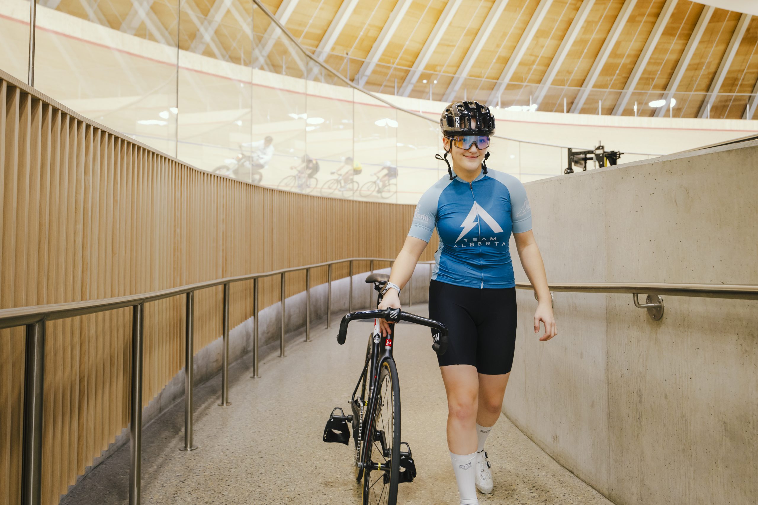 A teen girl, wearing a helmet, a Team Alberta bike jersey and shorts, walks her bike up a curved concrete ramp inside a velodrome.