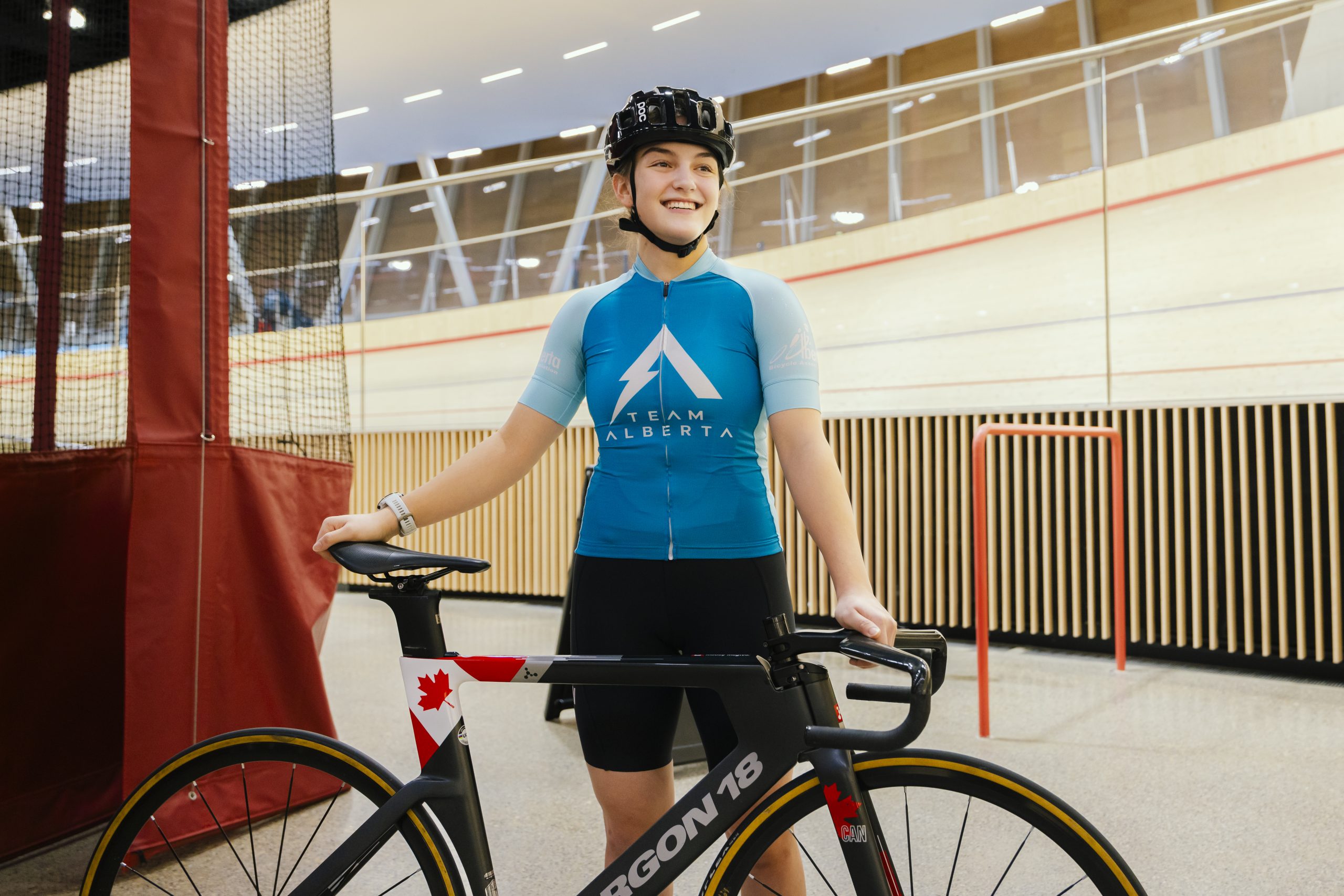 A teen girl, wearing a helmet, a Team Alberta bike jersey and shorts, holds her bike in the infield of an indoor cycling track.