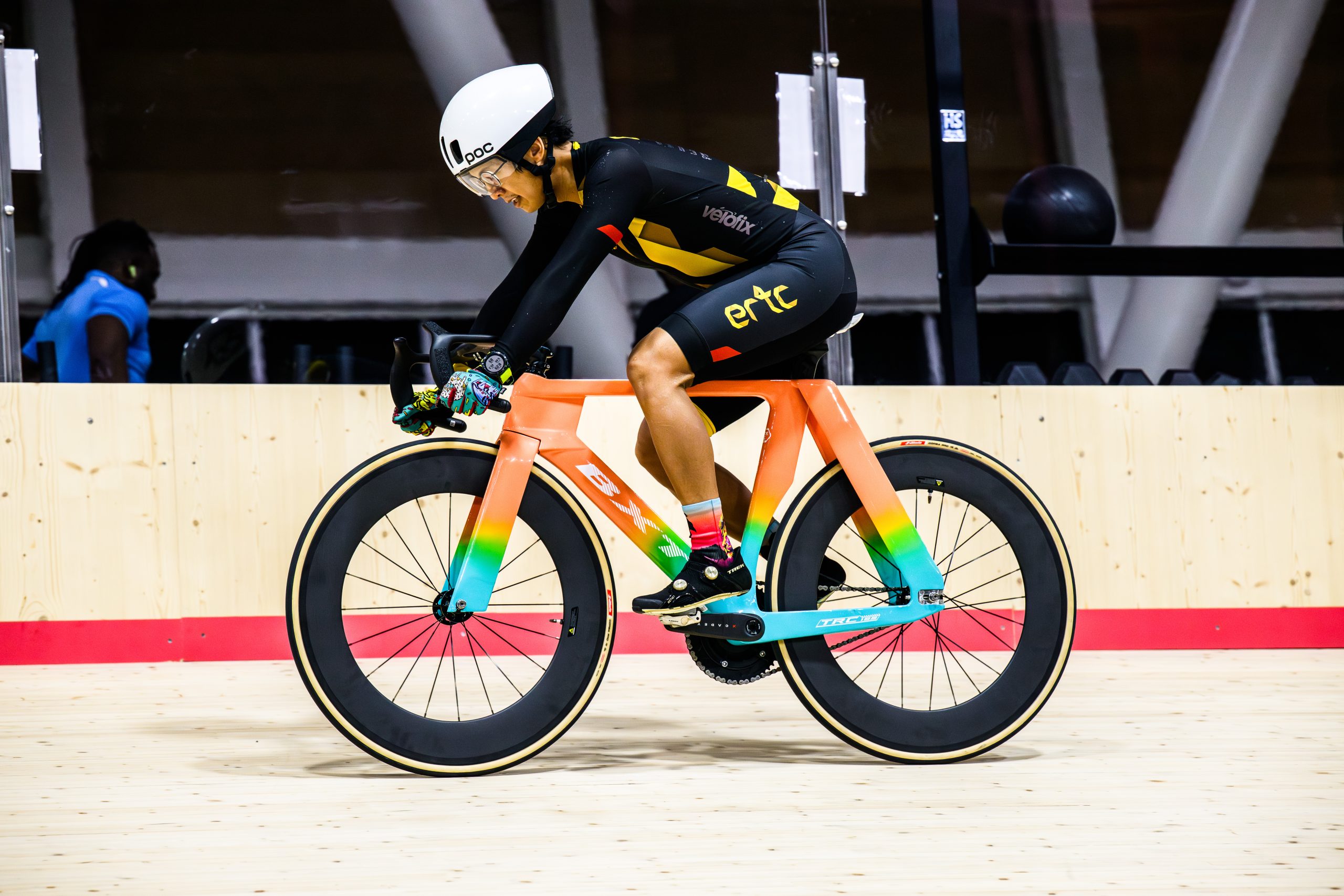 A cyclist, wearing a helmet, glasses, gloves and a skin suit, rides a bike on the indoor track of a velodrome.