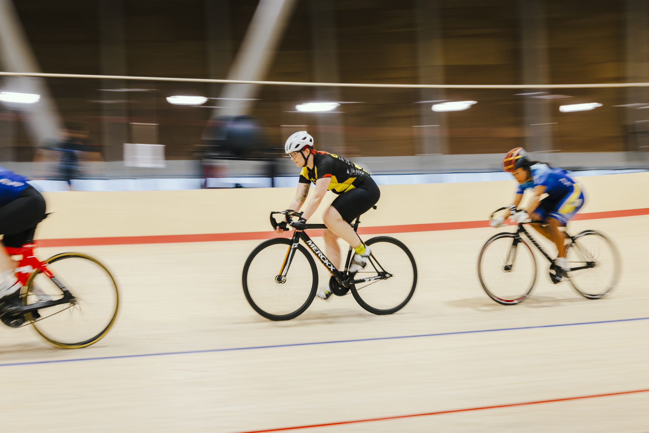 Three cyclists race in single file on a wooden track in an indoor velodrome.