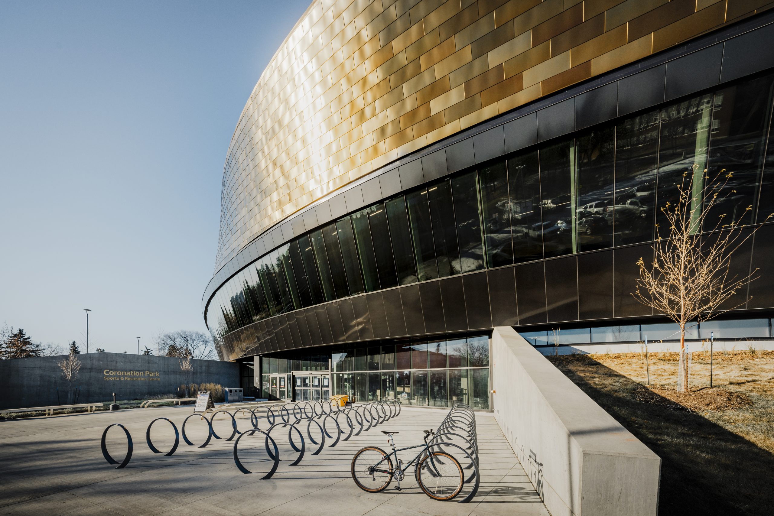A bike, locked to a circular bike rack, stands in front of an oval-shaped building covered with windows and coppery tiles.