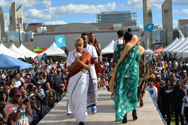 Black people displaying cultural fashions on a catwalk in front of a large crowd as part of an outdoor festival. CR_Africanival