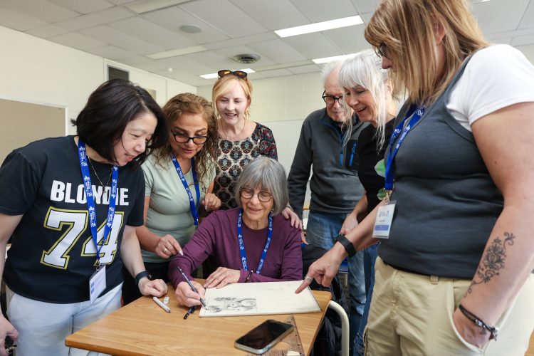 Five women and one man stand around a woman sitting at a desk, drawing on a pad of paper.