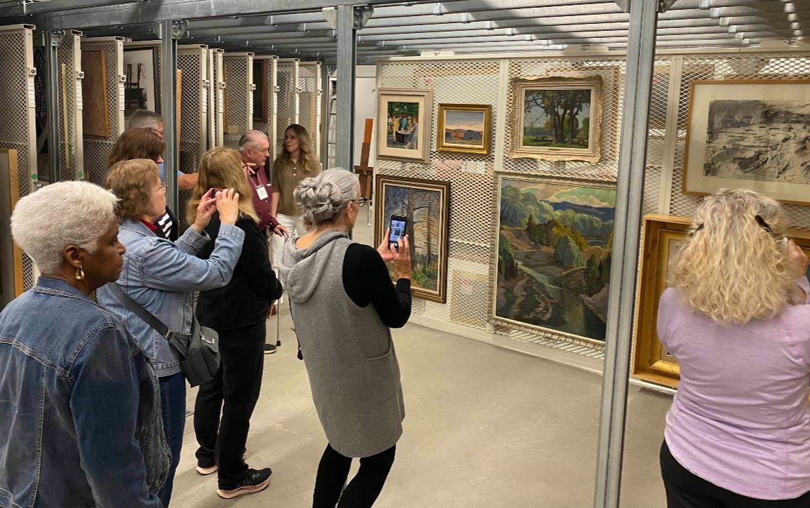 A group of students point their smart phones at a wall of framed paintings.