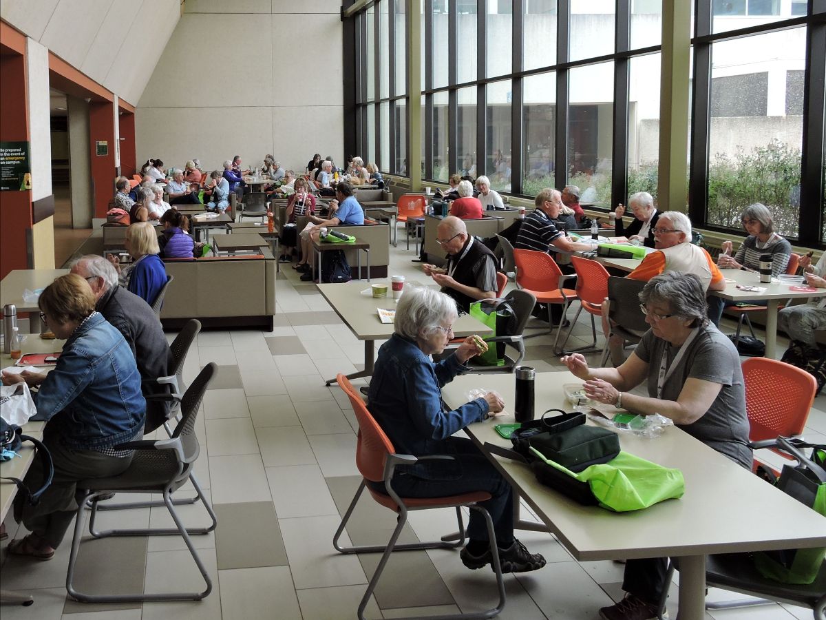 Men and women sit at tables in an cafeteria with a wall of large windows.