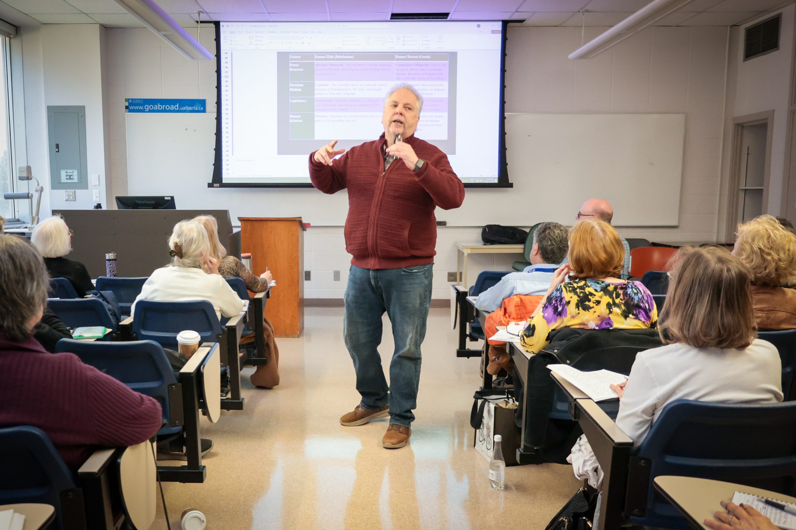 A man walks and talks in the middle of a classroom, as adult students sit in rows of seats on either side of him.