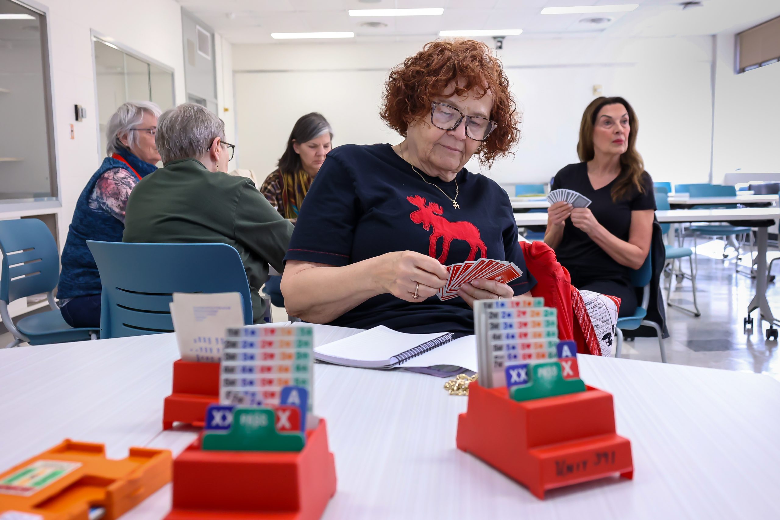 A woman sits a table, looking down at the playing cards in her hands, as a group of four women play cards at a table behind her.