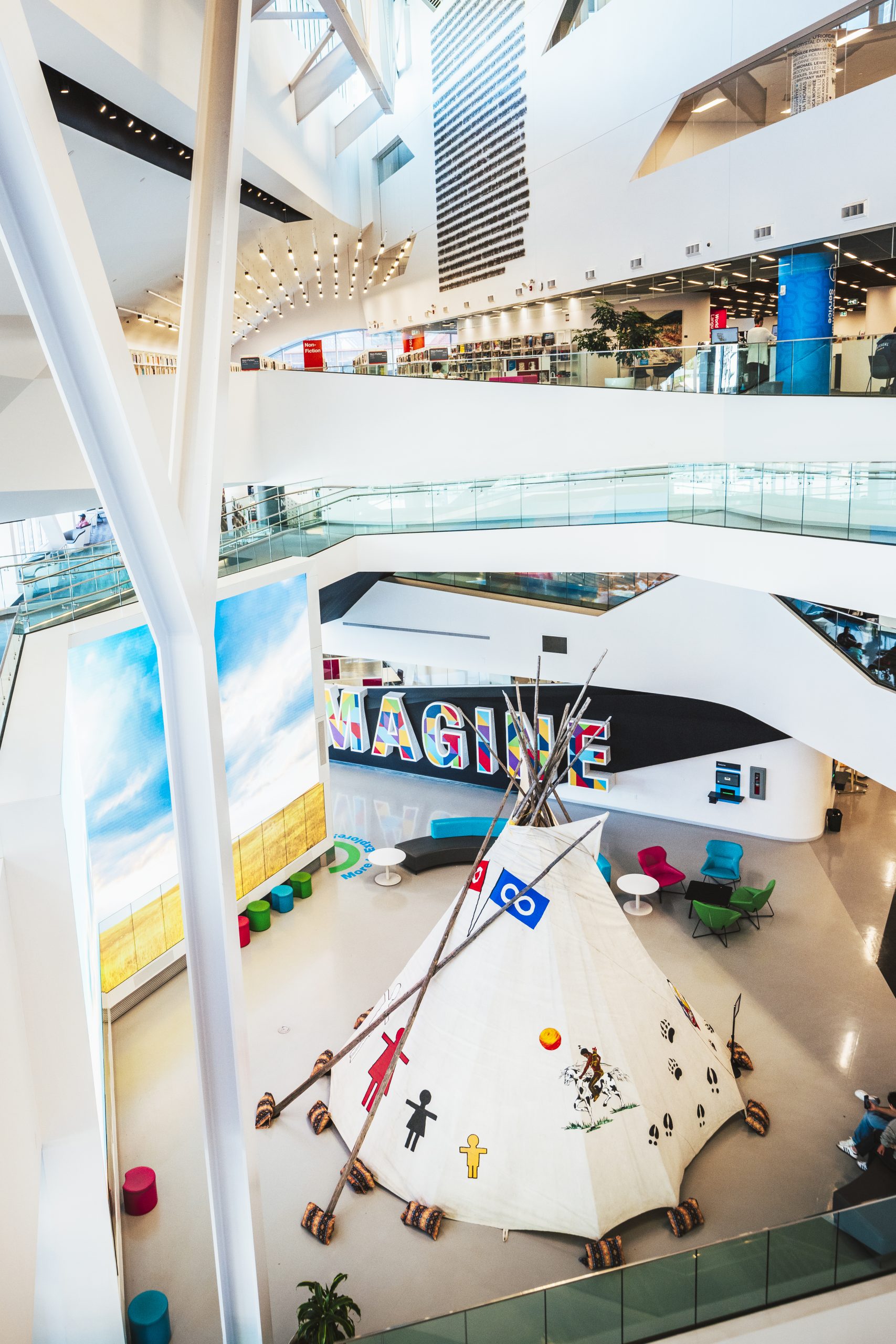 A teepee, surrounded by chairs and stools, stands on the main floor of a library.