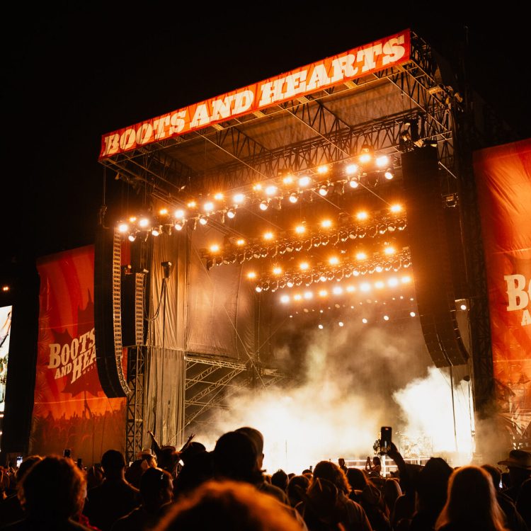 A crowd watching an outdoor music festival stage at night with lights and smoke.