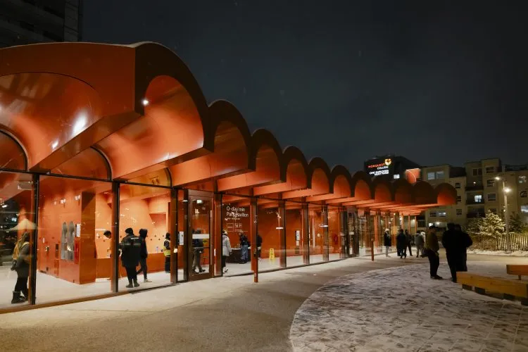 People walk inside and outside a glassed-walled building with a multi-arched red roof on a winter evening.