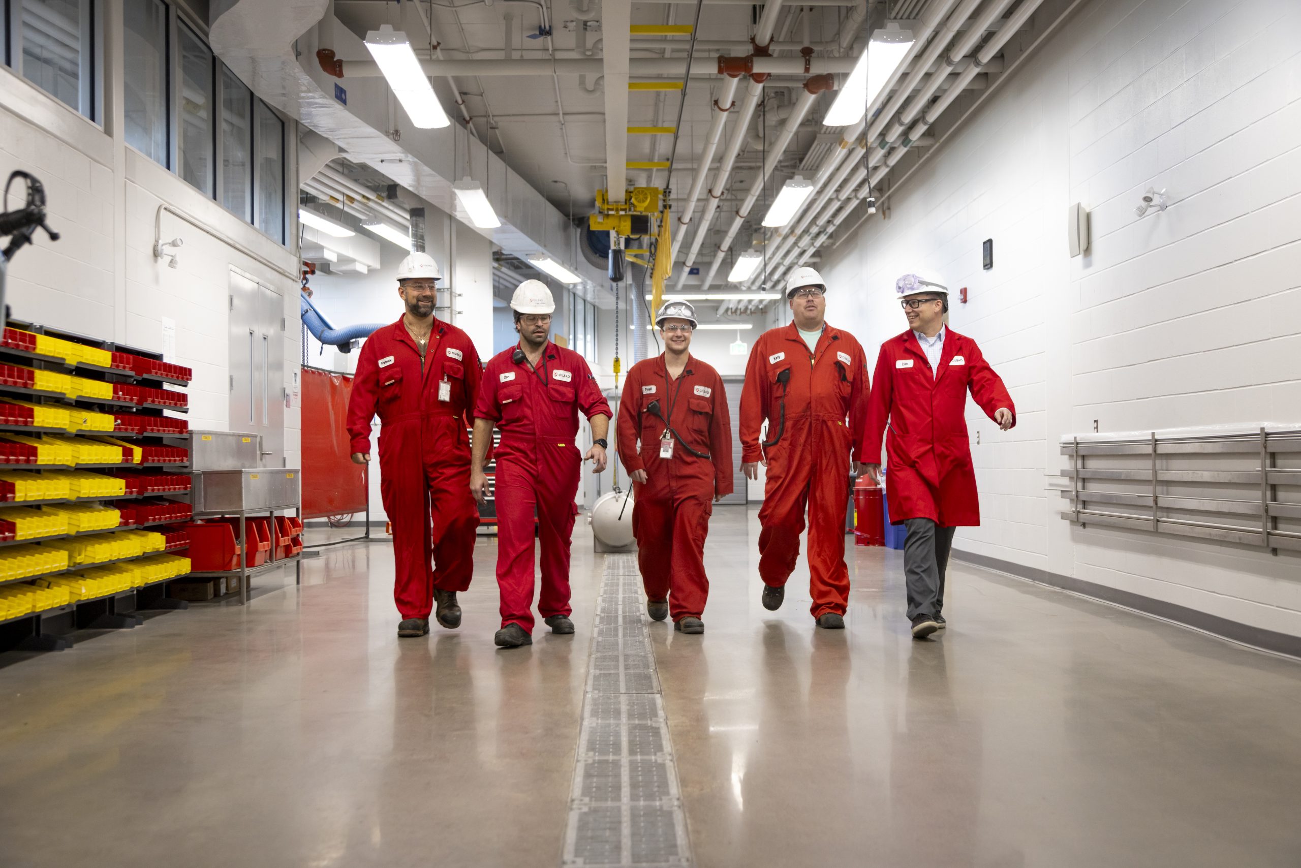 Five team members walking down a large industrial hallway in red jumpsuits and white hard hats.