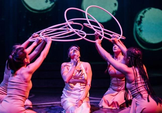 Five women performing on stage, kneeling in a circle holding small hoops and bathed in pink light. 