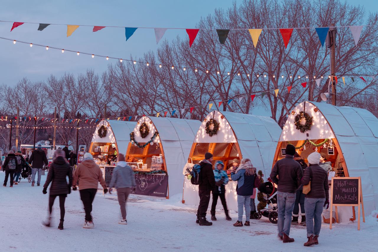 People walking and shopping at an outdoor holiday market with festive lit up booths.
