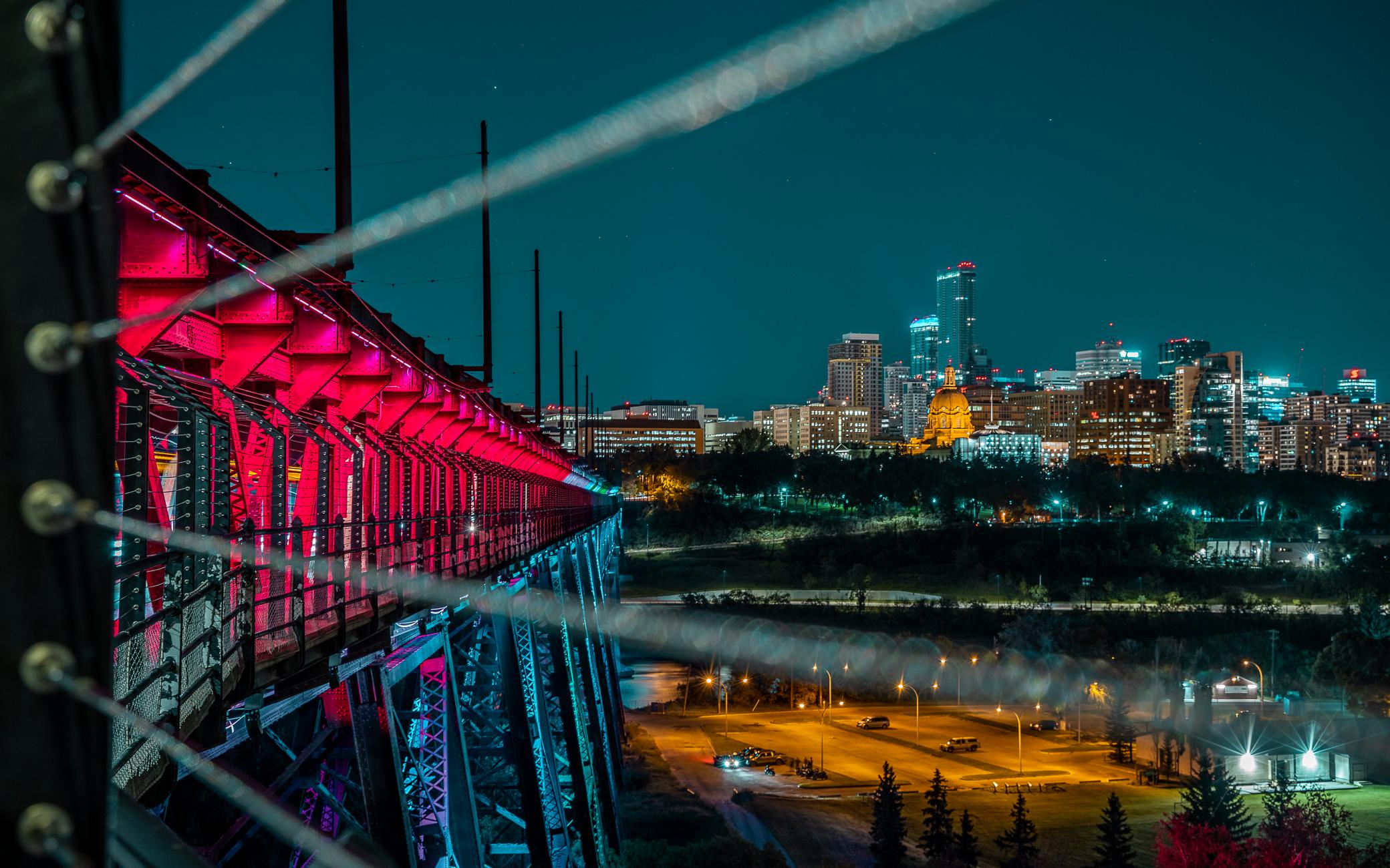 A close up of the High Level Bridge lit in red and blue with the city skyline at night.