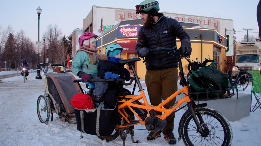 A dad holds his orange bike on a snowy sidewalk and looks at his two daughters sitting in a bike seat behind his own. They are all wearing bike helmets, gloves and winter parkas.